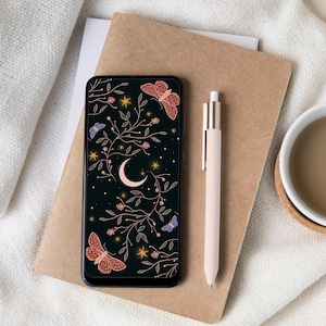 May include: A black smartphone displays a celestial design with a crescent moon, butterflies, stars, and floral elements. A beige pen rests beside the phone on a brown notebook. A white mug sits on a coaster in the corner, with a white cloth in the background.