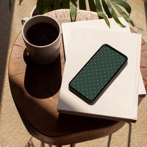 May include: A smartphone with a dark green polka dot design lies on a stack of books. A mug of coffee sits on a wooden table. A plant is visible in the background.