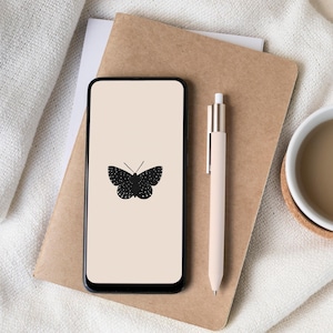 Könnte beinhalten: Ein Smartphone zeigt eine schwarze Schmetterlingsgrafik auf beigem Hintergrund. Ein beiger Stift liegt neben dem Telefon auf einem braunen Notizbuch. Eine Tasse Kaffee steht auf einem Untersetzer neben einem weißen Tuch.