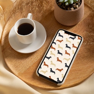 May include: A smartphone with a white case displays a repeating pattern of dachshund dogs in black, brown, and tan against a cream background. A white coffee cup and saucer sit on a wooden tray, next to a small potted plant.