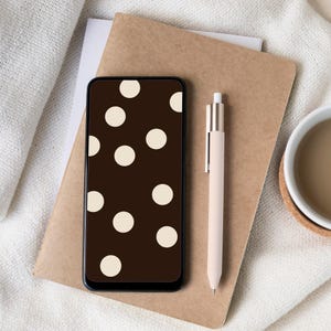 May include: A smartphone with a brown polka dot pattern on the screen rests on a brown notebook. A beige pen and a cup of coffee sit nearby on a white cloth. The scene is a top-down view, creating a minimalist aesthetic.