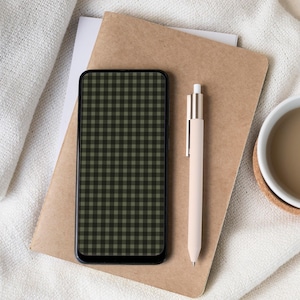 May include: A smartphone with a green and black checkered pattern screen rests on a brown notebook. A beige pen and a white mug with a brown liquid are also present. The items are arranged on a white, textured surface.