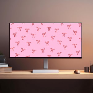 May include: A computer monitor displays a pink background with a repeating pattern of red bows. The monitor sits on a wooden desk with a mouse, pens, and a stack of books. The overall aesthetic is clean and modern.