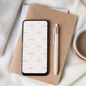 May include: A smartphone with a bow pattern screen, a beige pen, a brown notebook, and a cup of coffee on a white cloth. The phone's screen displays a repeating pattern of light blue bows and dots on a pink grid.