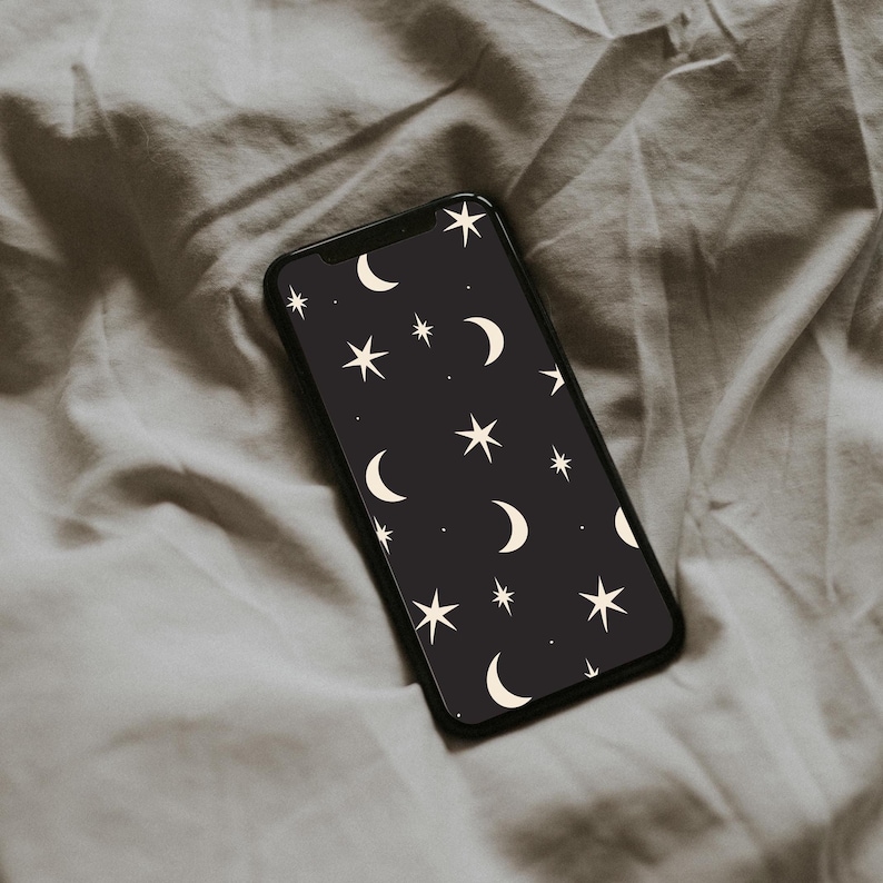 May include: A black smartphone displays a celestial pattern of white crescent moons and stars against a dark background. The phone is positioned on a wrinkled, light gray fabric, creating a soft, textured backdrop.