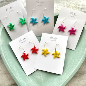 May include: Six pairs of colorful, handmade polymer clay earrings shaped like pinwheels. Each pair is displayed on a white card with the text "Ella Rae OXFORD".