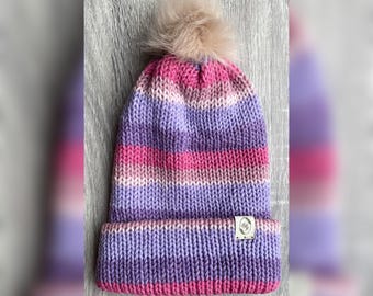 Puede incluir: Un gorro tejido a mano en tonos de rosa, morado y blanco con un pompón de piel sintética. El gorro presenta rayas horizontales y un borde doblado. Una pequeña etiqueta blanca está cosida en el borde.