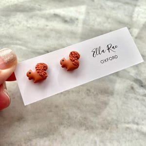 Könnte beinhalten: Ein Paar kleine, orangebraune Eichhörnchen-Ohrstecker. Die Eichhörnchen sind aus Polymer Clay gefertigt und haben detaillierte Merkmale. Die Ohrringe werden auf einer weißen Karte mit dem Text "Ella Rae Oxford" präsentiert.