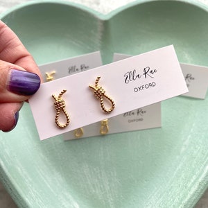 Puede incluir: Un par de pendientes de nudo dorados en una tarjeta blanca con el texto "Ella Rae Oxford".
