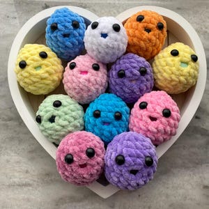 May include: A collection of 11 colorful crocheted balls with happy faces. Each ball is a different color, including yellow, pink, blue, green, purple, and orange. The balls are arranged in a heart shape on a white surface.