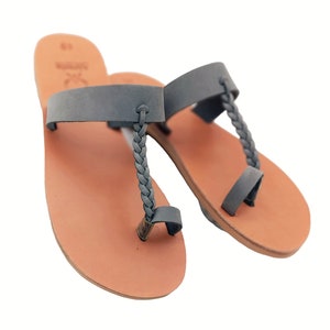 May include: A pair of gray leather sandals with a braided toe strap and a strap across the top of the foot. The sandals have a light brown sole and are set against a white background. The sandals are a size 9.