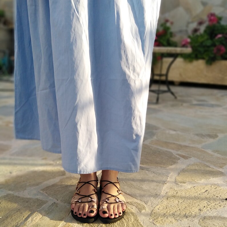 K&ouml;nnte beinhalten: Ein Paar schwarze Ledersandalen mit goldenen Akzenten und gekreuzten Riemen. Die Sandalen werden von einer Person in einem hellblauen Kleid getragen.