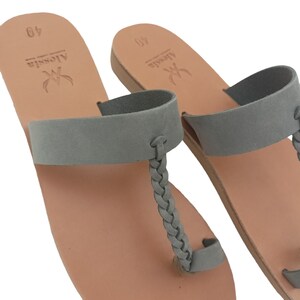 May include: A pair of gray leather sandals with a braided toe strap and a wide strap across the top of the foot. The sandals have a light brown sole and the brand name "Alessia" is stamped on the insole.