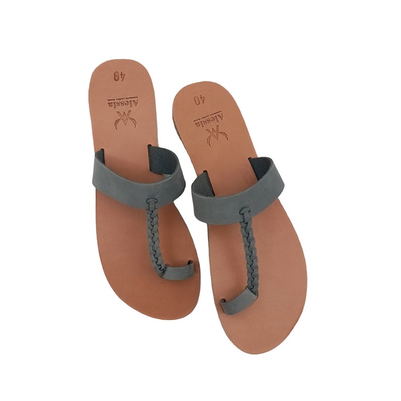 May include: A pair of gray leather sandals with a braided toe strap and a wide strap across the top of the foot. The sandals have a brown footbed and are labeled with the brand name "Alessia" and size 40.