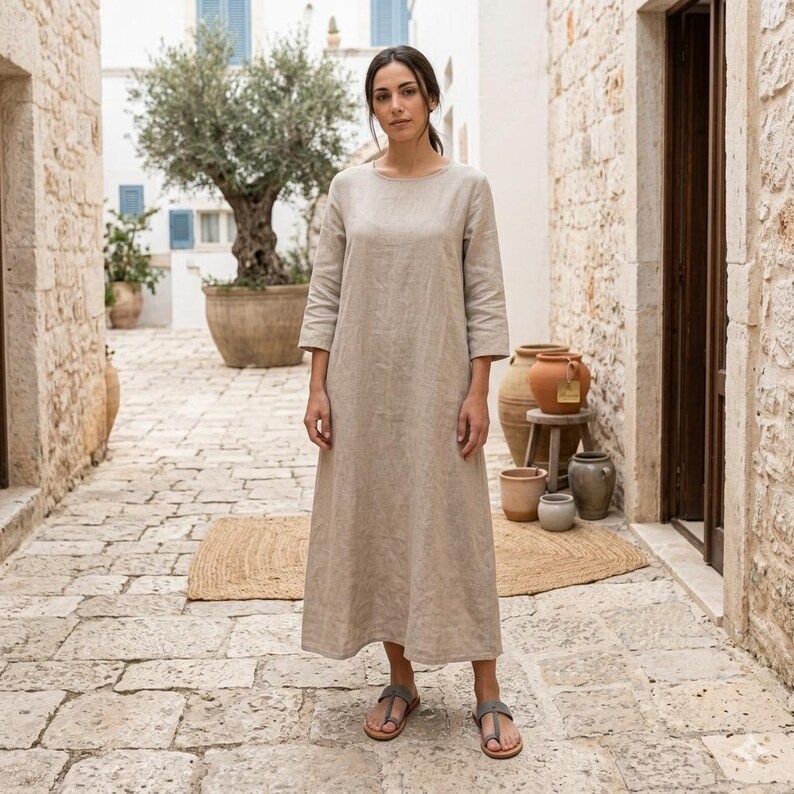 May include: A beige linen dress with a round neckline and three-quarter sleeves. The dress is ankle-length and worn with gray sandals. The setting is an outdoor stone walkway with a large tree and pottery.
