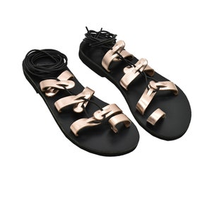 K&ouml;nnte beinhalten: Ein Paar schwarze Ledersandalen mit ros&eacute;goldenen Riemen und Schnallen. Die Sandalen haben eine flache Sohle und eine Zehenschlaufe.