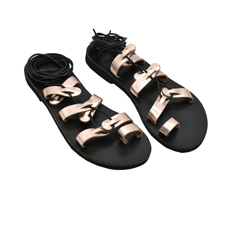K&ouml;nnte beinhalten: Ein Paar schwarze Ledersandalen mit ros&eacute;goldenen Riemen und Schnallen. Die Sandalen haben eine flache Sohle und eine Zehenschlaufe.