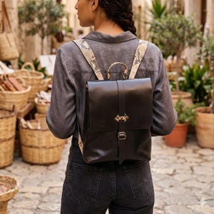May include: A dark brown leather backpack featuring a flap closure with a vertical strap and a metal buckle. The backpack has adjustable shoulder straps with a beige and brown snakeskin pattern and a small top handle for carrying.