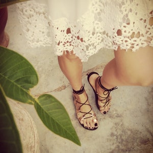 K&ouml;nnte beinhalten: Ein Paar schwarze Ledersandalen mit Schn&uuml;rsenkeln, die am Bein hochgebunden werden. Die Sandalen haben eine goldene Metallic-Oberfl&auml;che und werden von einer Person mit einem wei&szlig;en Spitzenrock getragen.