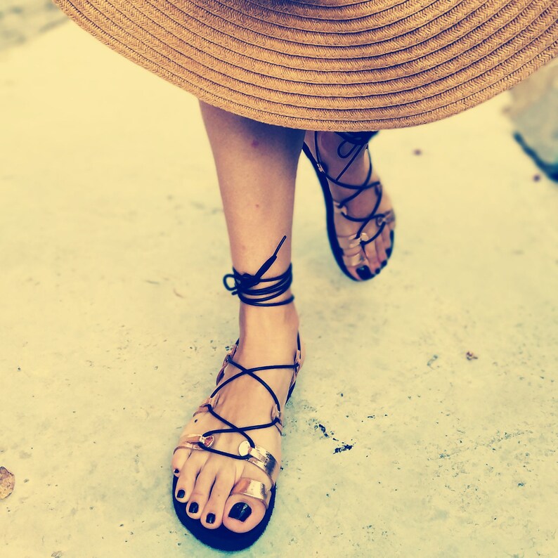 K&ouml;nnte beinhalten: Ein Paar goldene Sandalen mit schwarzen Schn&uuml;rsenkeln, die bis zu den Beinen gebunden sind. Die Sandalen haben eine runde Zehenpartie und werden an den F&uuml;&szlig;en getragen.