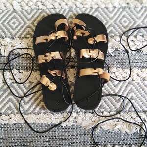 K&ouml;nnte beinhalten: Schwarze Ledersandalen mit ros&eacute;goldenen Riemen und Schn&uuml;ren. Die Sandalen haben einen Gladiator-Stil mit mehreren Riemen, die sich um den Kn&ouml;chel wickeln.