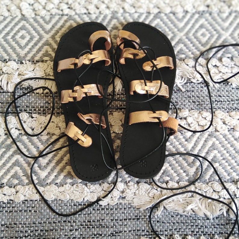 K&ouml;nnte beinhalten: Schwarze Ledersandalen mit ros&eacute;goldenen Riemen und Schn&uuml;ren. Die Sandalen haben einen Gladiator-Stil mit mehreren Riemen, die sich um den Kn&ouml;chel wickeln.