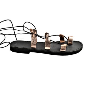 K&ouml;nnte beinhalten: Schwarze Ledersandalen mit ros&eacute;goldenen Riemen und Schn&uuml;ren. Die Sandalen haben eine flache Sohle und sind am Bein gebunden.