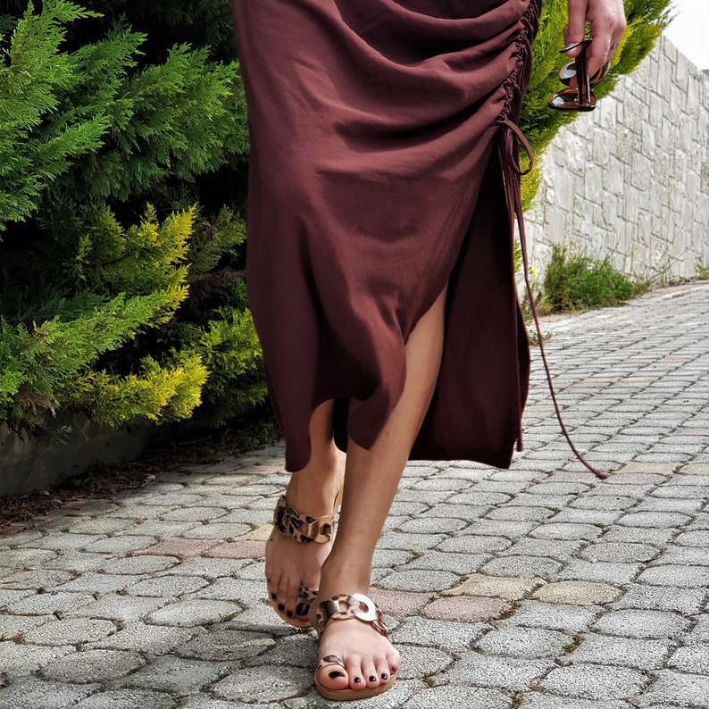 May include: A person wearing a dark burgundy dress with a side slit and drawstring detail. Gold-toned sandals with a leopard print design are worn on the feet. The person is walking on a brick-paved pathway.