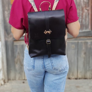 Könnte beinhalten: Ein schwarzer Lederrucksack mit einem Schlangenhautmusterriemen. Der Rucksack hat eine Klappe mit einem Schnallenverschluss und ein kleines Fach auf der Vorderseite.