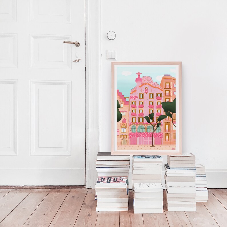 May include: A framed illustration of a pink building with a whimsical design, surrounded by trees and flowers. The building has a unique architectural style with balconies and windows. The illustration is set against a blue sky with white clouds.
