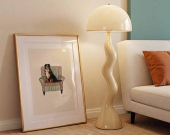 May include: A framed artwork featuring a dog sitting in a striped armchair, next to a cream-colored, wavy-stemmed floor lamp with a mushroom-shaped shade. A magazine is open on the floor.