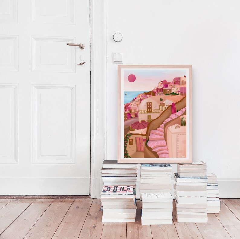 May include: A framed illustration of a pink and white village with a pink sun in the sky. The village is built on a hillside with a winding staircase leading up to the top. The illustration is on a shelf with a stack of books.