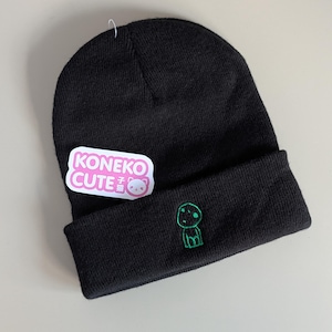 May include: A black knit beanie featuring a small green embroidered character on the cuff. A white rectangular sticker with pink text reading KONEKO CUTE and a small cat icon is attached to the side of the beanie with a thin plastic fastener.