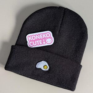 May include: A dark charcoal gray knit beanie featuring a folded cuff with an embroidered sunny-side-up egg patch. A rectangular white sticker with pink text reading KONEKO CUTE and a small cat icon is attached to the upper portion of the beanie.