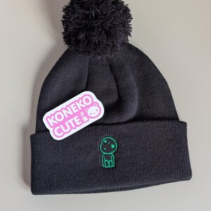 May include: A black knit beanie featuring a large black pom-pom on top and a folded cuff. The cuff displays a small green embroidered character graphic and a white sticker with pink text that reads KONEKO CUTE with a small cat icon.