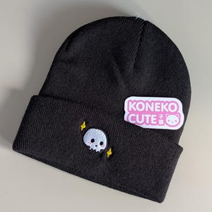May include: A black knit beanie featuring an embroidered white skull with pink cheeks and two yellow four-pointed stars. A white rectangular sticker with pink text reading KONEKO CUTE and a small cat face icon is attached to the folded cuff.