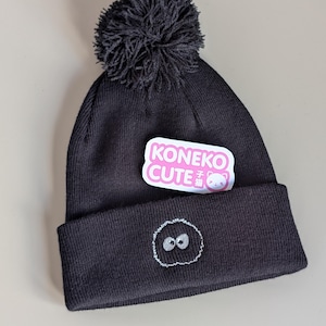 May include: A black knit beanie featuring a dark gray pom-pom on top and a folded cuff. The cuff displays an embroidered circular gray creature with two white eyes. A white sticker with pink text reading KONEKO CUTE is attached to the beanie.