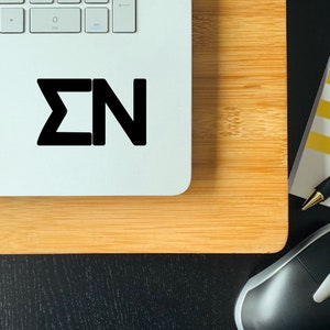 May include: A white laptop with a black sticker that says "&Sigma;&Nu;" on the top of the laptop. The laptop is sitting on a wooden surface.