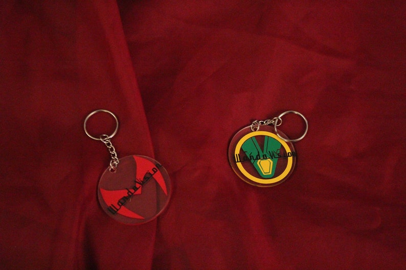 Puede incluir: Dos llaveros redondos con anillos de metal. El llavero de la izquierda tiene un dise&ntilde;o rojo y negro con el texto "WandaVision" en blanco. El llavero de la derecha tiene un dise&ntilde;o amarillo, verde y rojo con el texto "WandaVision" en negro.