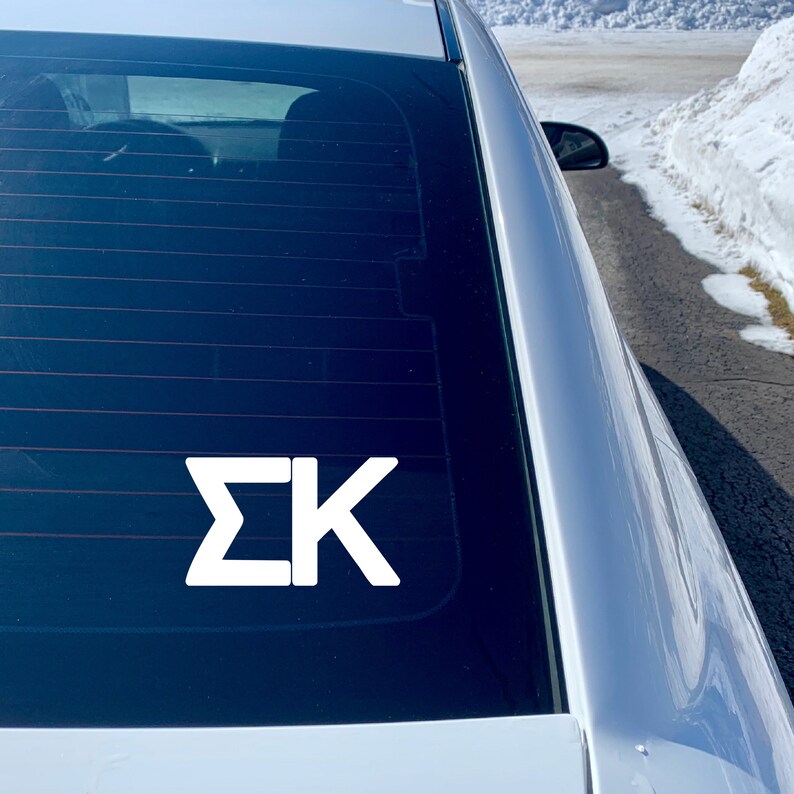 May include: White vinyl decal with the letters "&Sigma;K" on a black car window.