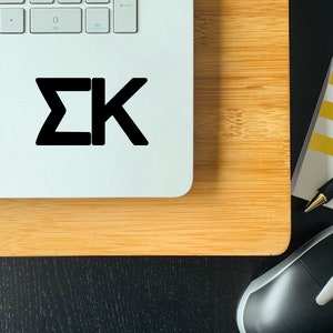 May include: A white laptop with a black "&Sigma;&Kappa;" sticker on the top. The laptop is sitting on a wooden surface. A black computer mouse is in the lower right corner of the image.