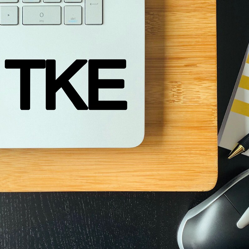 May include: A laptop computer with a black sticker that says "TKE" on the top of the case. The laptop is sitting on a wooden surface.