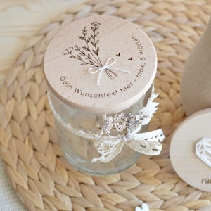 Könnte beinhalten: Ein Holzdeckel mit einem Blumenmuster und dem Text "Dein Wunschtext hier - max. 5 Wörter" befindet sich auf einem durchsichtigen Glasgefäß. Das Gefäß ist mit einem weißen Spitzenband und getrockneten Blumen verziert.