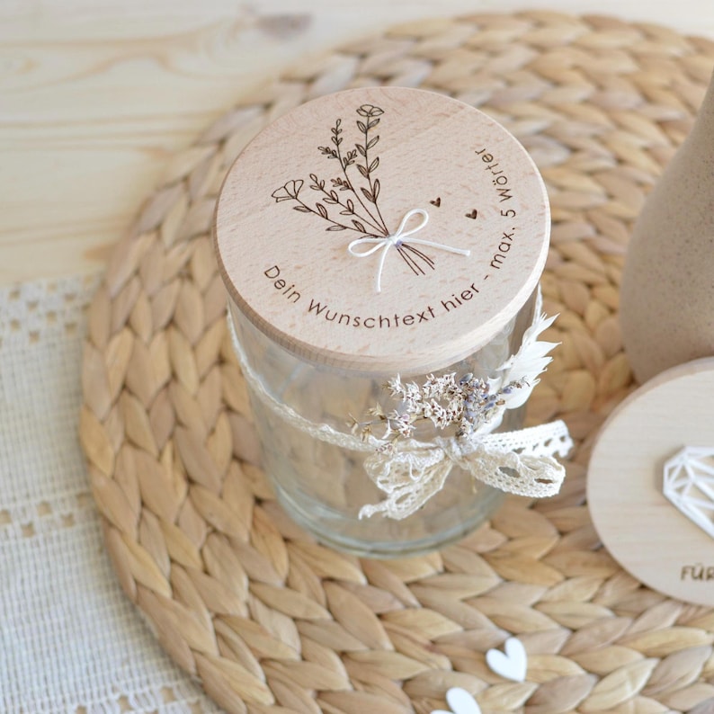 Könnte beinhalten: Ein Holzdeckel mit einem Blumenmuster und dem Text "Dein Wunschtext hier - max. 5 Wörter" befindet sich auf einem durchsichtigen Glasgefäß. Das Gefäß ist mit einem weißen Spitzenband und getrockneten Blumen verziert.