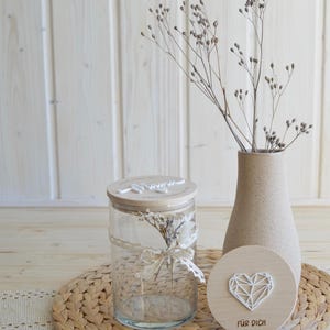 May include: A glass jar with a wooden lid and a small bouquet of dried flowers inside. The jar is sitting on a woven placemat with a wooden heart-shaped sign that says "F&uuml;r Dich" in German.