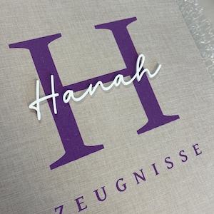 Pode incluir: Uma pasta revestida de tecido bege com uma letra H grande de cor roxa impressa no centro. O nome Hanah est&aacute; escrito em letra cursiva branca sobre a letra H, e a palavra ZEUGNISSE est&aacute; impressa em letras mai&uacute;sculas roxas abaixo.
