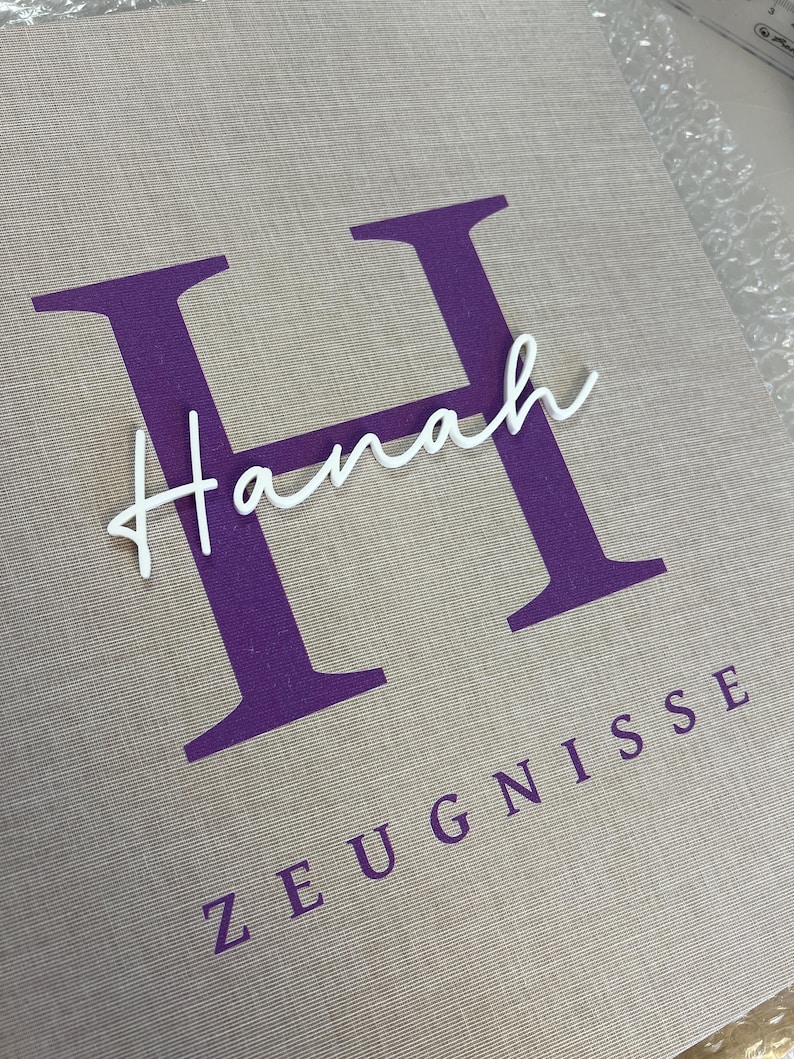 Pode incluir: Uma pasta revestida de tecido bege com uma letra H grande de cor roxa impressa no centro. O nome Hanah est&aacute; escrito em letra cursiva branca sobre a letra H, e a palavra ZEUGNISSE est&aacute; impressa em letras mai&uacute;sculas roxas abaixo.