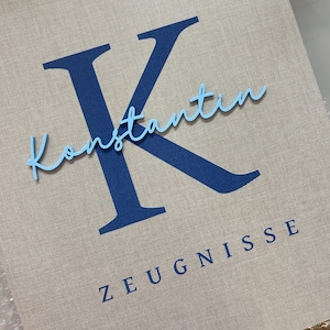 Pode incluir: Uma pasta revestida de tecido bege com uma letra K grande em azul escuro, o nome Konstantin em letra cursiva azul claro e a palavra ZEUGNISSE em letras mai&uacute;sculas azul escuro impressas na capa frontal.