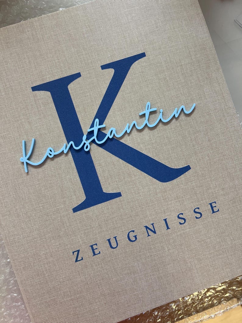 Pode incluir: Uma pasta revestida de tecido bege com uma letra K grande em azul escuro, o nome Konstantin em letra cursiva azul claro e a palavra ZEUGNISSE em letras mai&uacute;sculas azul escuro impressas na capa frontal.
