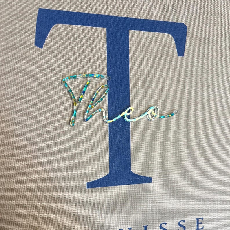 Pode incluir: Uma letra T mai&uacute;scula grande em azul escuro impressa num fundo de tecido bege. Sobreposta &agrave; letra est&aacute; uma etiqueta com o nome Theo em letra cursiva, feita de um material transparente com pequenas part&iacute;culas de purpurina multicolorida.
